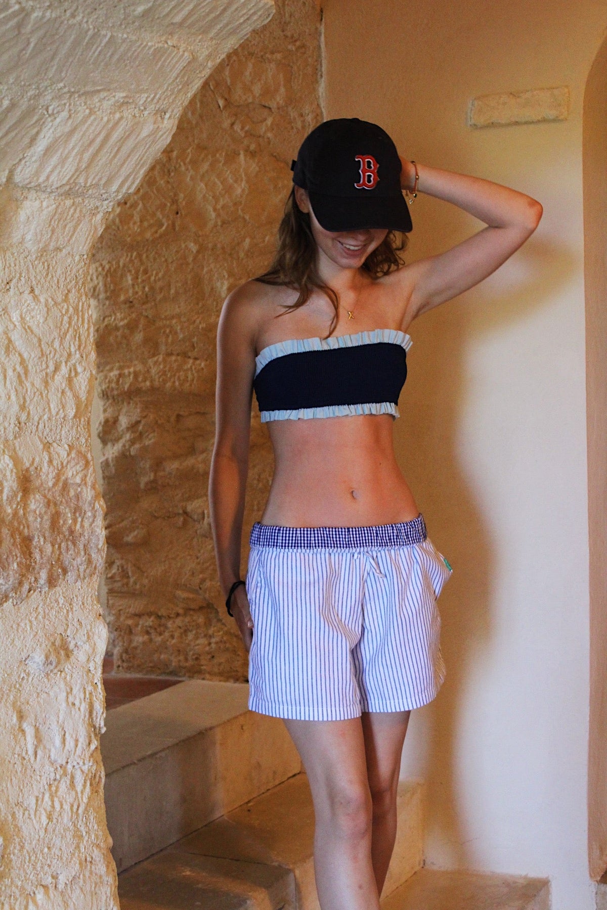 Woman wearing a black and white striped top and shorts with a baseball cap in a stone-walled room.