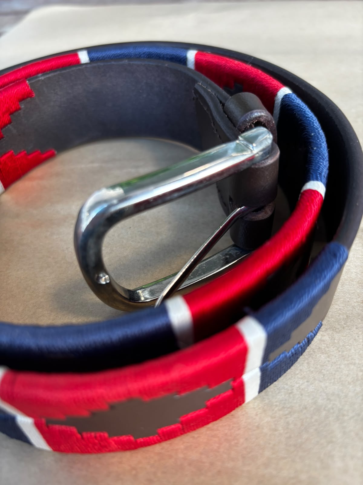 A close-up of a woven belt with a red, white, and blue pattern, a dark gray center, and a silver metal buckle, coiled on a neutral surface.