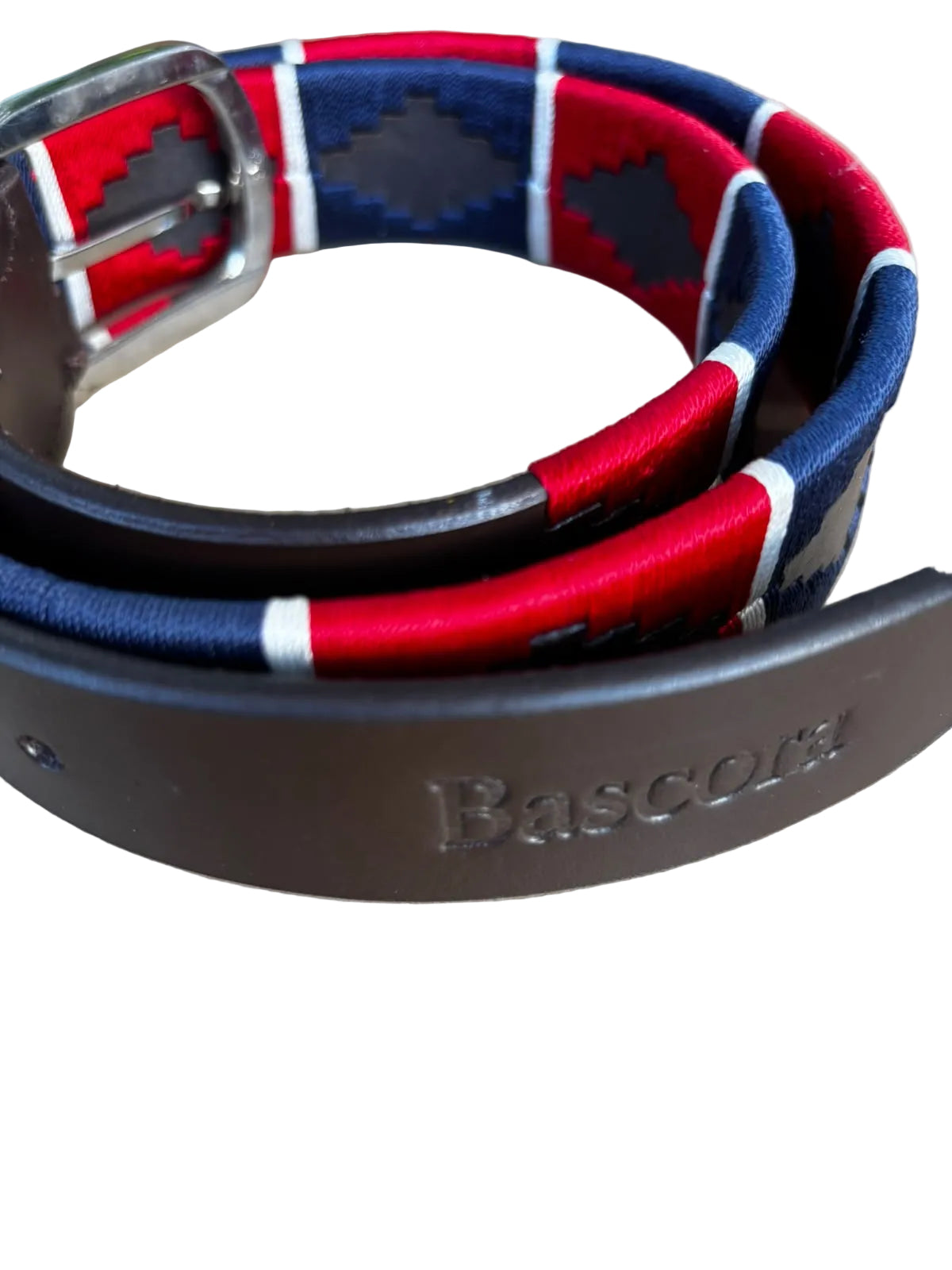 A close-up of a Bascora brand belt with a dark brown leather strap and a woven fabric inlay featuring red, navy blue, and white diamond patterns, resting on a neutral beige surface.