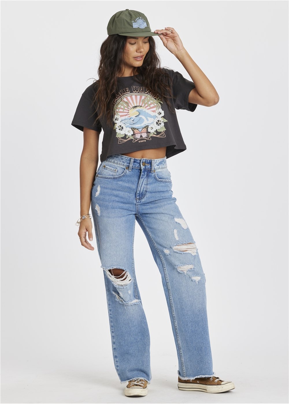 Woman wearing a graphic t-shirt and distressed jeans on a white background touching her hat